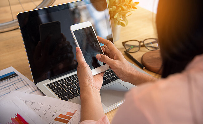 Eine Frau sitzt an einem Schreibtisch mit einem Laptop und hält ein Smartphone in der Hand